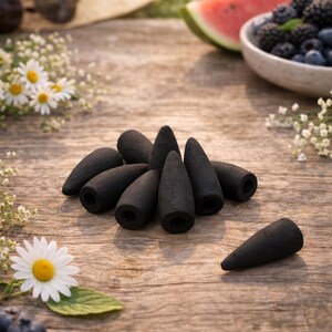 May include: A group of black incense cones on a weathered wooden surface. The cones are accompanied by daisies, blueberries, blackberries, and a slice of watermelon. A straw hat and sunglasses are in the background.