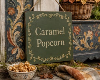 Może przedstawiać: Kwadratowy zielony drewniany szyld z jutowym sznurkiem, zawierający napis Caramel Popcorn kremowymi literami, otoczony malowaną bordiurą z liści. Ceramiczna miska wypełniona popcornem karmelowym stoi na zwietrzałej drewnianej powierzchni.