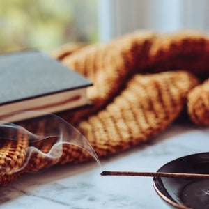 May include: A smoldering incense stick with smoke rising above it, resting on a black dish. The stick is on a white marble surface with a brown knitted blanket in the background.
