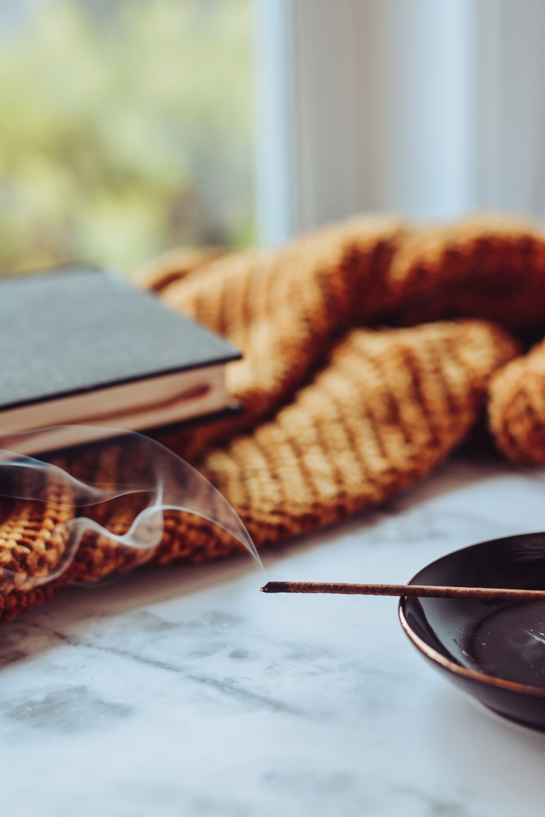 May include: A smoldering incense stick with smoke rising above it, resting on a black dish. The stick is on a white marble surface with a brown knitted blanket in the background.