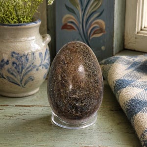 May include: A polished, egg-shaped stone sculpture featuring a mottled brown, black, and grey granite-like texture. The object is displayed in an upright position, resting securely on a small, clear, circular acrylic stand for stability and display.