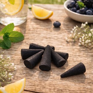 May include: Several black incense cones are displayed on a wooden table, with a glass of water containing lemon and mint, a bowl of blueberries, and lemon wedges. The setting is outdoors with natural light.