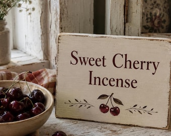 Może przedstawiać: Rustykalny drewniany szyld z napisem "Sweet Cherry Incense" w ciemnoczerwonych literach. Szyld przedstawia ilustrację wiśni i stoi obok miski ze świeżymi wiśniami. W tle bukiet białych kwiatów.