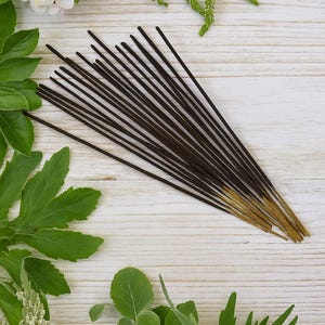 May include: A collection of dark brown incense sticks with golden tips, arranged on a light wooden surface. Green leaves and white flowers surround the incense, creating a natural and aromatic theme. The incense sticks are a popular item for aromatherapy and meditation.