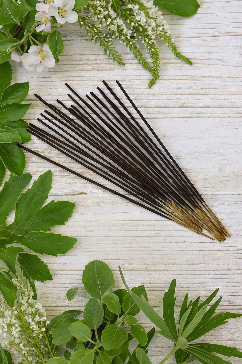 May include: A collection of dark brown incense sticks with golden tips, arranged on a light wooden surface. Green leaves and white flowers surround the incense, creating a natural and aromatic theme. The incense sticks are a popular item for aromatherapy and meditation.