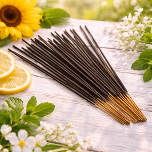 May include: A collection of dark brown incense sticks with gold tips, presented on a white wooden surface. The image features a yellow sunflower, lemon slices, and sprigs of white flowers and green leaves, suggesting a natural, aromatic product.