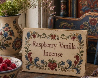 以下が含まれることがあります： 「Raspberry Vanilla Incense」とバーガンディ色の文字が書かれた素朴な木製看板。赤、青、緑の花柄の縁取りが施されています。看板は使い古された木製のテーブルの上に置かれ、背景にはラズベリーのボウルと花の入った花瓶があります。