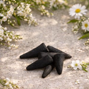 May include: A collection of black incense cones arranged on a textured, light-colored surface, surrounded by white flowers and greenery. The cones have a rough texture and a conical shape, suggesting a natural product.