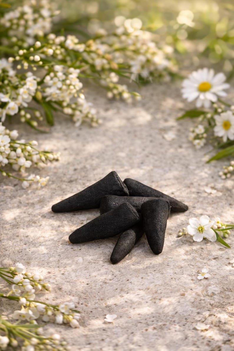 May include: A collection of black incense cones arranged on a textured, light-colored surface, surrounded by white flowers and greenery. The cones have a rough texture and a conical shape, suggesting a natural product.