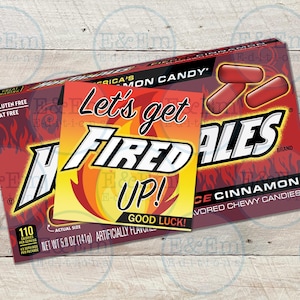 May include: A red, white, and orange box of Fireballs cinnamon candies. The box has a red and white flame design and the text "Let's get Fired Up!" on a separate sticker. The box is on a wooden surface.