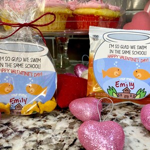 May include: A Valentine's Day treat bag with a goldfish bowl design. The bag is filled with goldfish crackers and has the text "I'm so glad we swim in the same school! Happy Valentine's Day!" and "Your friend Emily".