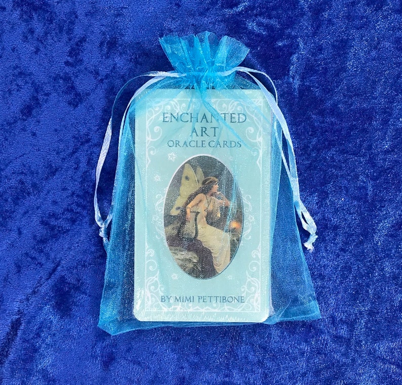 K&ouml;nnte beinhalten: Ein blaues und wei&szlig;es Orakelkarten-Deck mit dem Titel "Enchanted Art Oracle Cards" und dem Autor "By Mimi Pettibone" in einem hellblauen Kordelbeutel.