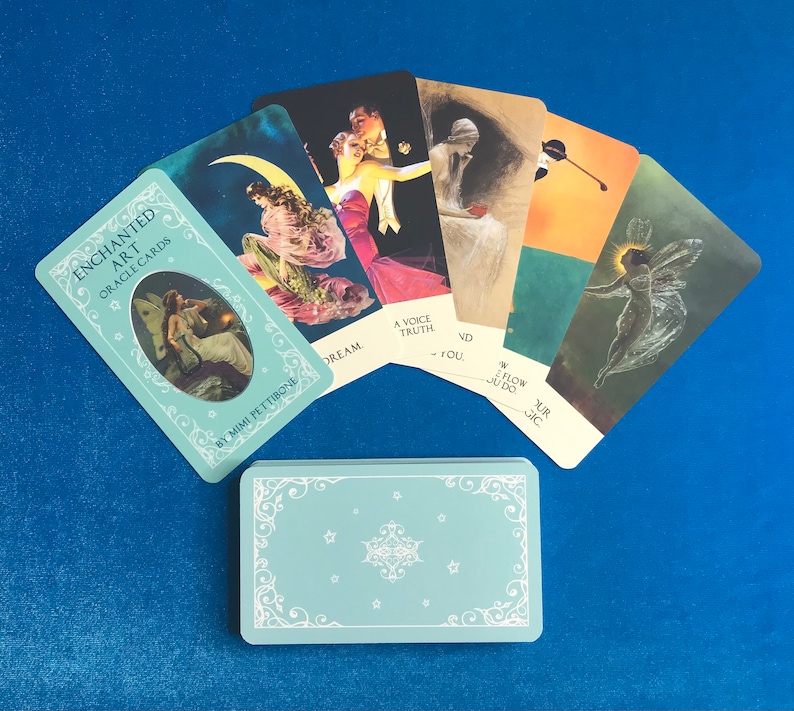 K&ouml;nnte beinhalten: Ein Set mit Enchanted Art Oracle Cards von Mimi Pettibone. Die Karten zeigen farbenfrohe Illustrationen und Texte wie "Dream", "A Voice Truth", "You", "Flow", "Your Magic" und "VD". Die Karten sind f&auml;cherf&ouml;rmig auf einer blauen Oberfl&auml;che angeordnet.