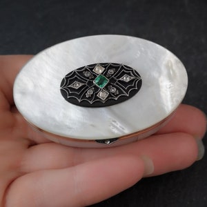 Op de afbeelding: Een wit parelmoer compact met een zwart emaille ovaal ontwerp met een groene smaragd en diamanten.