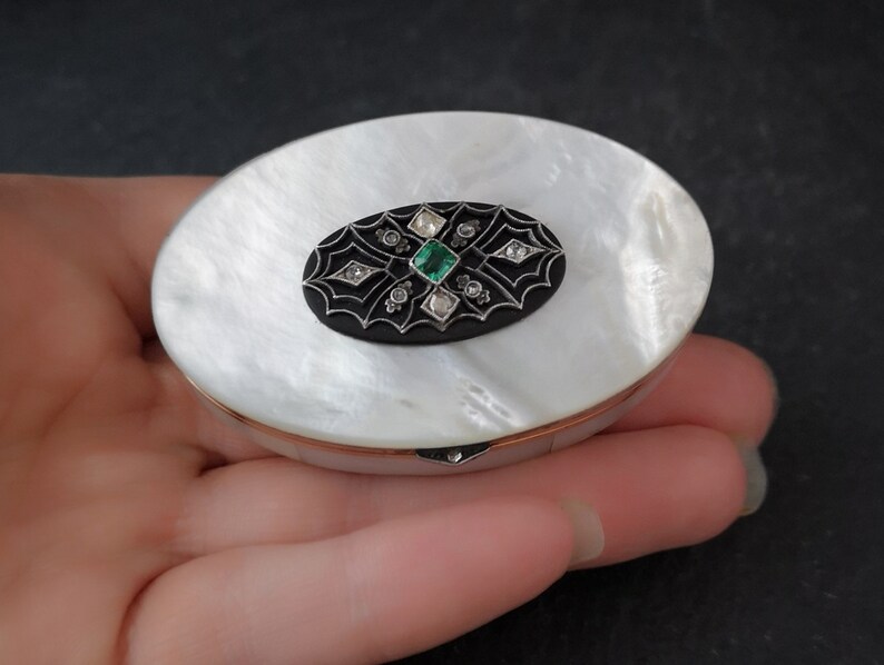 Op de afbeelding: Een wit parelmoer compact met een zwart emaille ovaal ontwerp met een groene smaragd en diamanten.