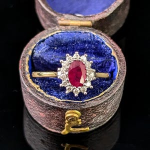 Peut inclure: Une bague en or ornée d'un rubis rouge profond de taille ovale, entouré d'un halo de petits diamants blancs ronds. La bague est présentée dans un petit écrin rond à la texture sombre, doublé d'un tissu en velours bleu vif.