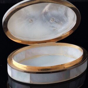Op de afbeelding: Een ovale, witte parelmoer sieraden doos met een gouden rand. De doos is open en laat een wit interieur zien.