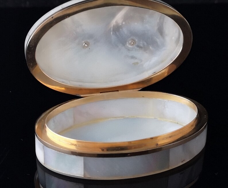 Op de afbeelding: Een ovale, witte parelmoer sieraden doos met een gouden rand. De doos is open en laat een wit interieur zien.