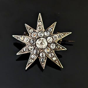 Op de afbeelding: Een stervormige broche met een centrale ronde heldere steen, omringd door meerdere kleinere heldere stenen in een metalen frame. Het ontwerp heeft acht puntige stralen die vanuit het midden uitlopen, elk bezet met heldere steentjes.