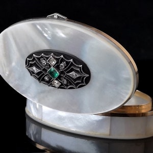 Op de afbeelding: Ovale nacre-sieradendoos met een scharnierend deksel. Het deksel heeft een zwart emaille ontwerp met een groene edelsteen en diamant accenten.