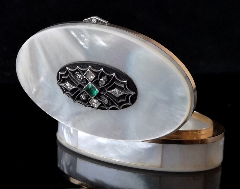 Op de afbeelding: Ovale nacre-sieradendoos met een scharnierend deksel. Het deksel heeft een zwart emaille ontwerp met een groene edelsteen en diamant accenten.