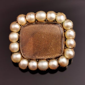 Può includere: Un fermaglio dorato con un pannello di vetro quadrato circondato da un bordo di perle bianche.