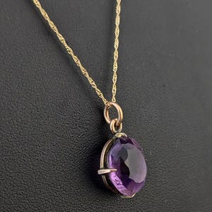 May include: A gold necklace featuring a thin, twisted chain and a pendant with a single, oval-shaped, faceted purple gemstone held in a metal setting with four prongs. The pendant hangs from a circular gold jump ring attached to the chain.