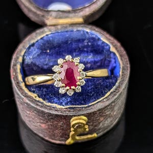 Peut inclure: Une bague en or ornée d'un rubis rouge taille ovale entouré d'un halo de petits diamants blancs ronds. La bague est présentée à l'intérieur d'un petit récipient cylindrique usé avec une doublure intérieure en velours bleu profond.