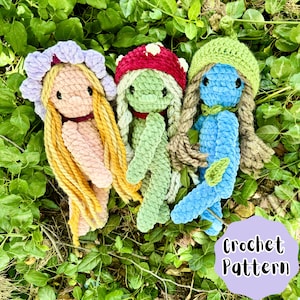 May include: Three crocheted dolls with different colored yarn and hair. The dolls are wearing different outfits. The doll on the left has a pink dress and yellow hair. The doll in the middle has a green dress and brown hair. The doll on the right has a blue dress and brown hair. The dolls are sitting on a bed of green grass.