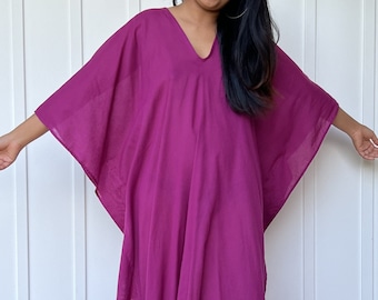 May include: A long, flowing magenta kaftan with a V-neckline. The kaftan is made of a lightweight fabric and has a relaxed fit. The model is wearing the kaftan with sandals. The text "MALA handworks Hollywood, MD" is visible on the bottom of the kaftan.