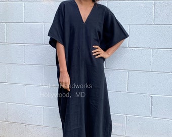 May include: A black, long-sleeved, v-neck kaftan dress with a relaxed fit. The dress is made of a soft, lightweight fabric and has a flowing silhouette. The model is wearing the dress with a pair of earrings. The dress is photographed against a white brick wall.  Mala Handworks Hollywood, MD