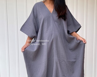 May include: A gray, V-neck caftan dress with wide sleeves. The dress is made of a textured fabric and falls to the ankles. The text "MAIA handworks Hollywood, MD" is visible on the garment.