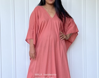 Puede incluir: Una larga kaftán de color rosa coral con cuello en V y mangas 3/4. La kaftán tiene una abertura lateral y está hecha de una tela ligera. El texto "MALA handworks Hollywood, MD" está impreso en la parte inferior de la kaftán.