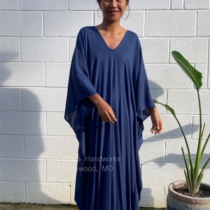 May include: A navy blue kaftan dress with a V-neckline. The dress is loose-fitting and has long sleeves. The model is standing in front of a white brick wall.
