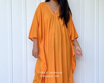 Puede incluir: Un vestido largo de caftán naranja fluido con escote en V. El vestido está hecho de una tela ligera y tiene un corte holgado. La modelo lleva el vestido con sandalias. MALA handworks Hollywood, MD