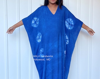 May include: A blue tie-dye kaftan with white circular patterns. The kaftan has a V-neck and long sleeves. The model is wearing the kaftan with black heeled sandals. The text "MALA handworks Hollywood, MD" is visible on the front of the kaftan.