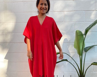 May include: A woman wearing a long, red, loose-fitting dress with a V-neck. The dress has short, wide sleeves. The woman is standing in front of a white brick wall with a potted plant to the right. The text "MaLa Handworks HOLLYWOOD MD" is visible on the dress.