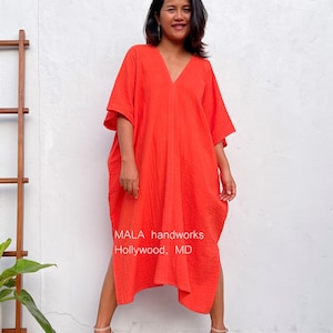 May include: A woman wearing a bright orange, loose-fitting, long-sleeved dress with a V-neck. The dress has side slits and a textured fabric. The woman is standing in front of a white wall with a wooden ladder and a potted plant. The text "MALA handworks Hollywood, MD" is visible at the bottom of the image.