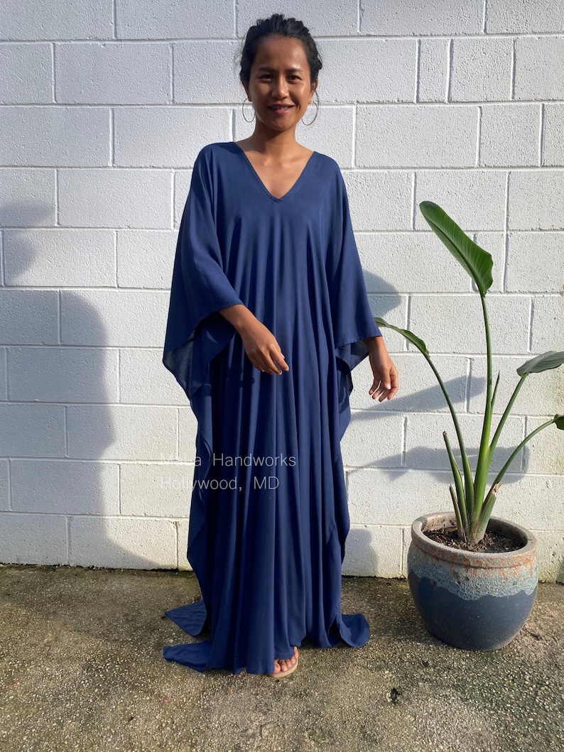 May include: A navy blue kaftan dress with a V-neckline. The dress is loose-fitting and has long sleeves. The model is standing in front of a white brick wall.