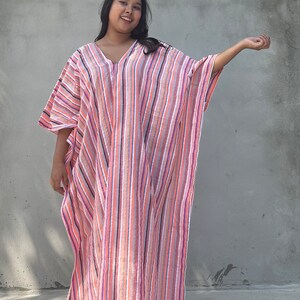 May include: A woman wearing a long, pink and white striped kaftan with a V-neckline. The kaftan has a relaxed fit and is made of a lightweight fabric.