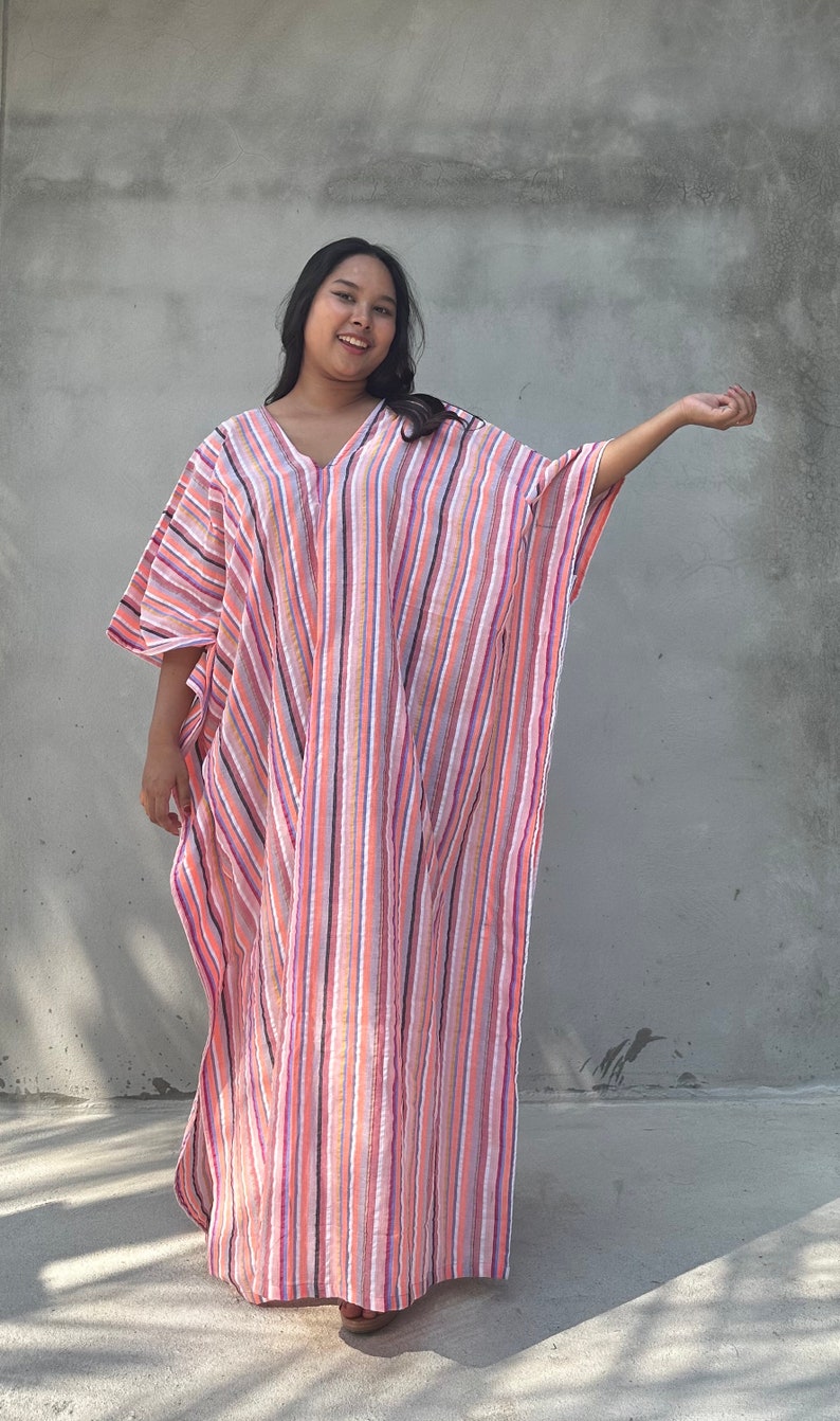 May include: A woman wearing a long, pink and white striped kaftan with a V-neckline. The kaftan has a relaxed fit and is made of a lightweight fabric.