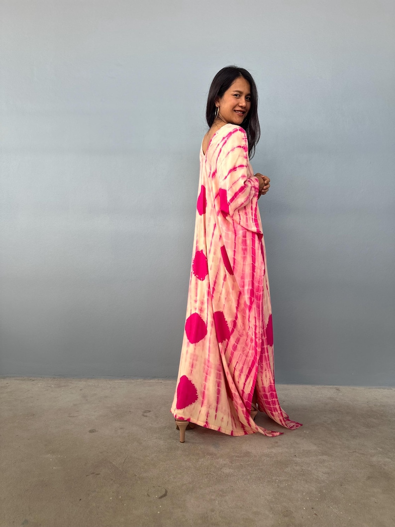 May include: A long, flowing dress with a tie-dye pattern in shades of pink and cream. The dress features large, circular pink accents and a V-neckline. The fabric drapes loosely, creating a relaxed silhouette.