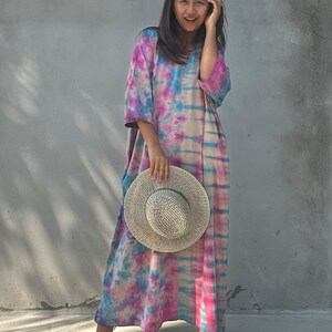 Op de afbeelding: Een vrouw draagt een lange, roze, blauwe en witte tie-dye jurk met een strohoed in haar hand.