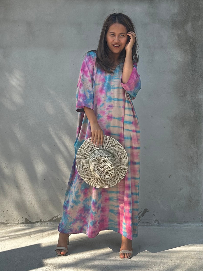 Op de afbeelding: Een vrouw draagt een lange, roze, blauwe en witte tie-dye jurk met een strohoed in haar hand.