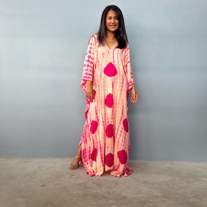 May include: A long, flowing dress with a V-neck and wide sleeves. The dress has a tie-dye pattern in pink and cream, with large, circular magenta accents. The dress is worn by a person standing in front of a grey wall.