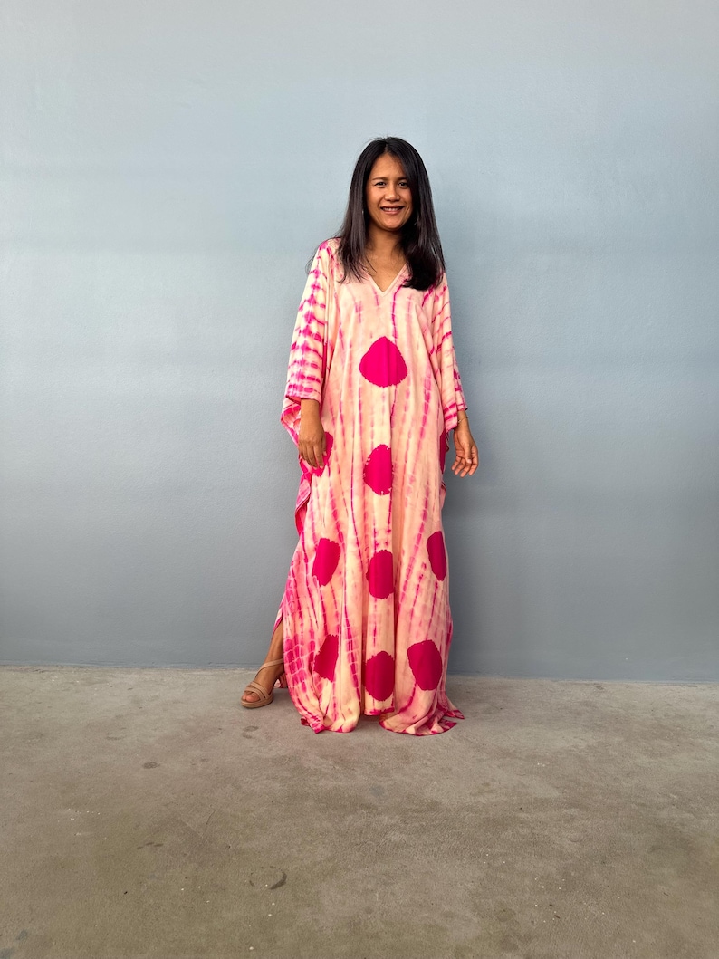 May include: A long, flowing dress with a V-neck and wide sleeves. The dress has a tie-dye pattern in pink and cream, with large, circular magenta accents. The dress is worn by a person standing in front of a grey wall.
