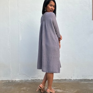 May include: A woman wearing a gray, loose-fitting, knee-length dress with a gathered neckline. She is standing in front of a white wall and wearing nude heels.