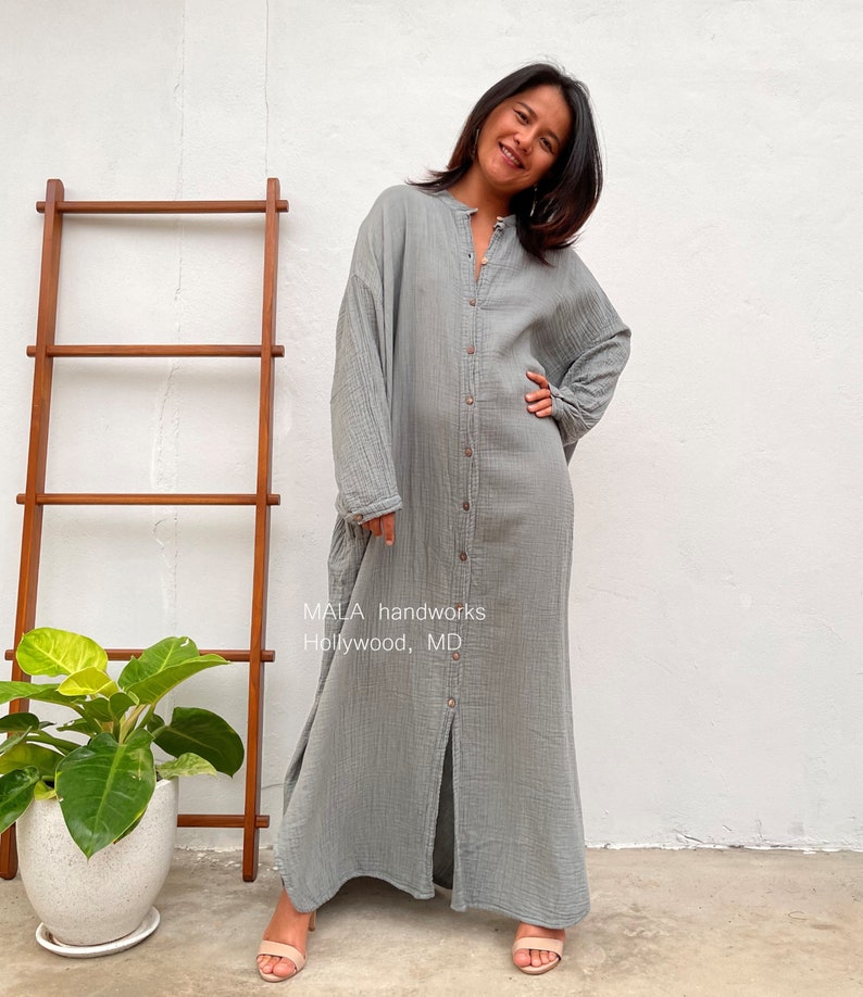 Pu&ograve; includere: Un lungo abito grigio chiaro con bottoni e una fessura sul davanti. L'abito ha una vestibilit&agrave; rilassata ed &egrave; realizzato in un tessuto morbido e testurizzato. La modella indossa l'abito con un paio di sandali color nude. Il testo "MALA handworks Hollywood, MD" &egrave; visibile sull'abito.