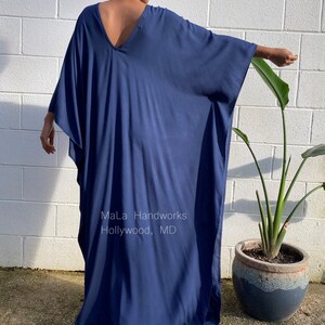 May include: A deep blue, long, flowing kaftan with a V-neckline. The kaftan is made of a soft, drapey fabric and has a relaxed fit. The model is wearing the kaftan with a simple hairstyle and no jewelry. The text "MaLa Handworks Hollywood, MD" is visible on the kaftan.