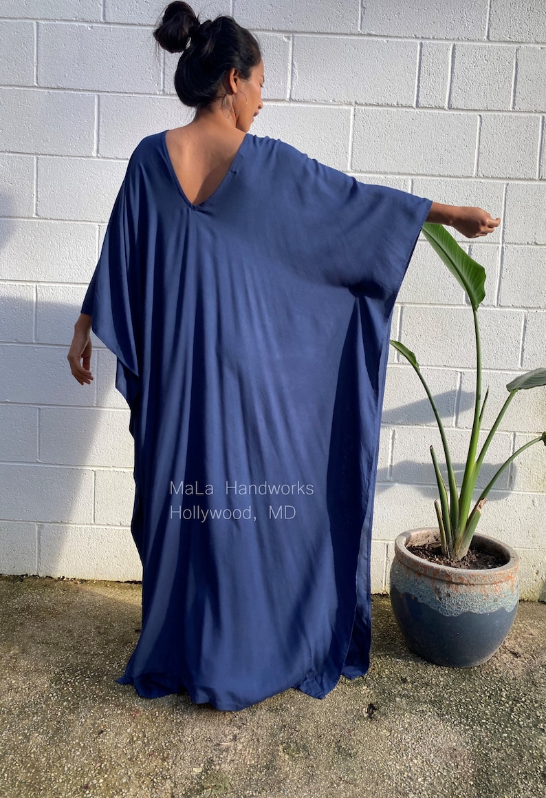 May include: A deep blue, long, flowing kaftan with a V-neckline. The kaftan is made of a soft, drapey fabric and has a relaxed fit. The model is wearing the kaftan with a simple hairstyle and no jewelry. The text "MaLa Handworks Hollywood, MD" is visible on the kaftan.
